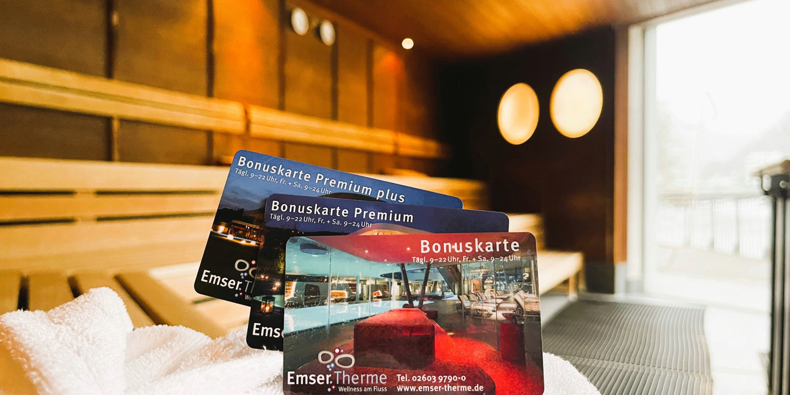 Drei Bonuskarten der Emser Therme liegen auf einem weißen Handtuch in einer Sauna. Der Hintergrund zeigt Holzverkleidung und große runde Fenster, die Licht hereinlassen.