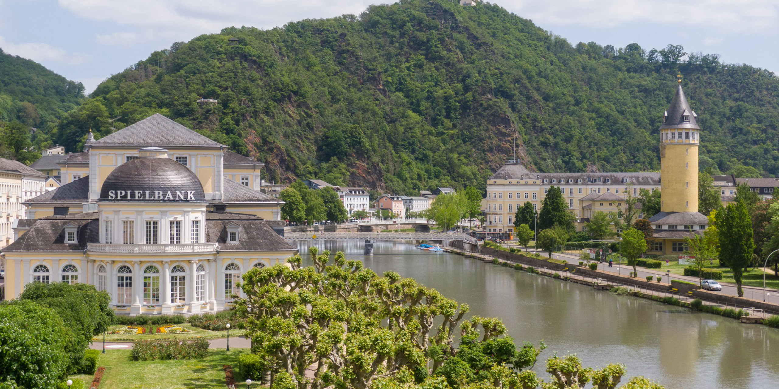 Panoramablick auf die Spielbank Bad Ems mit ihrer eleganten Fassade, umgeben von grünen Anlagen und dem ruhigen Fluss. Im Hintergrund erheben sich sanfte Hügel und historische Gebäude.