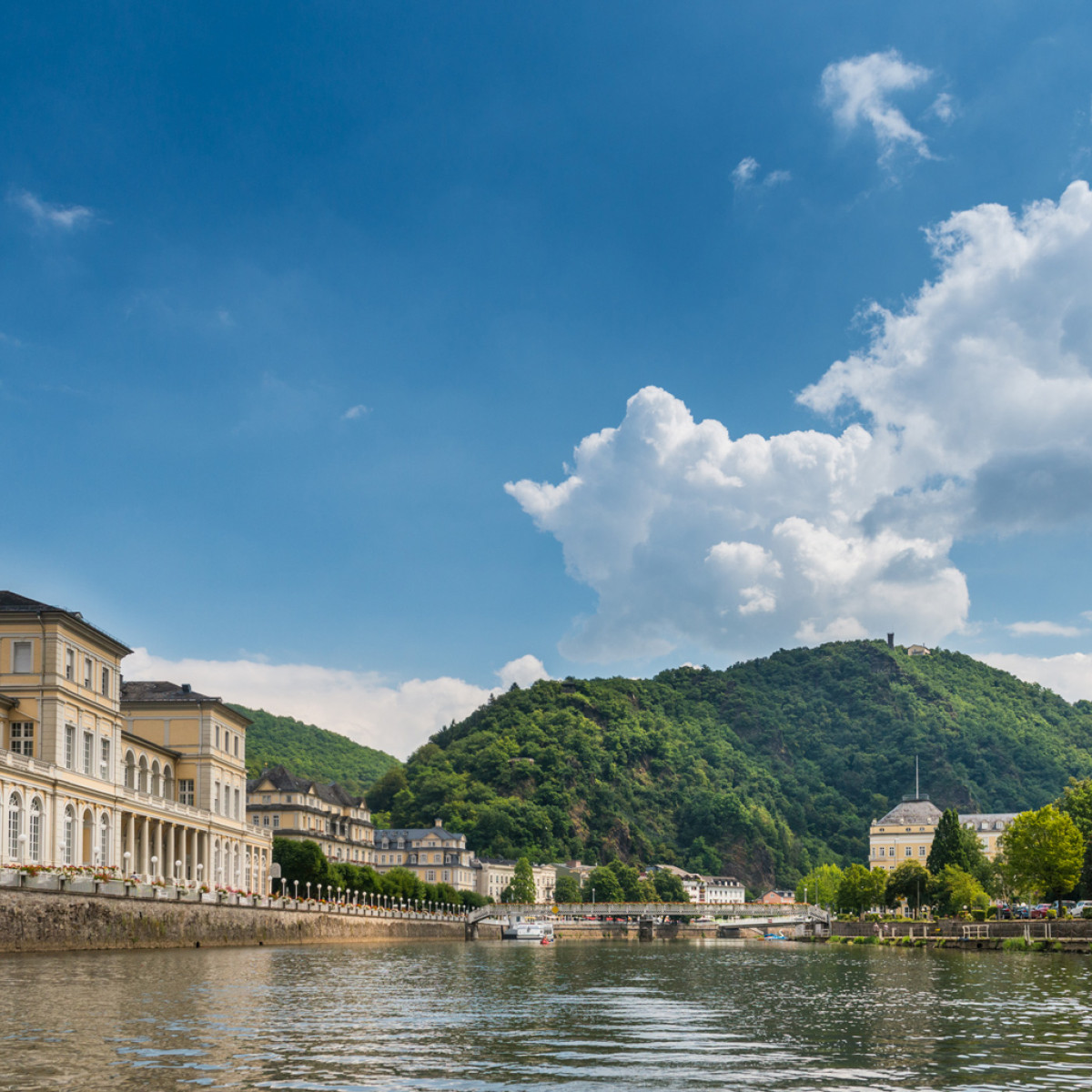 Panoramablick auf einen Fluss, umgeben von historischen Gebäuden und bewaldeten Hügeln. Ein Turm mit spitzem Dach steht in der rechten Bildhälfte, während weiße Wolken am blauen Himmel schweben.