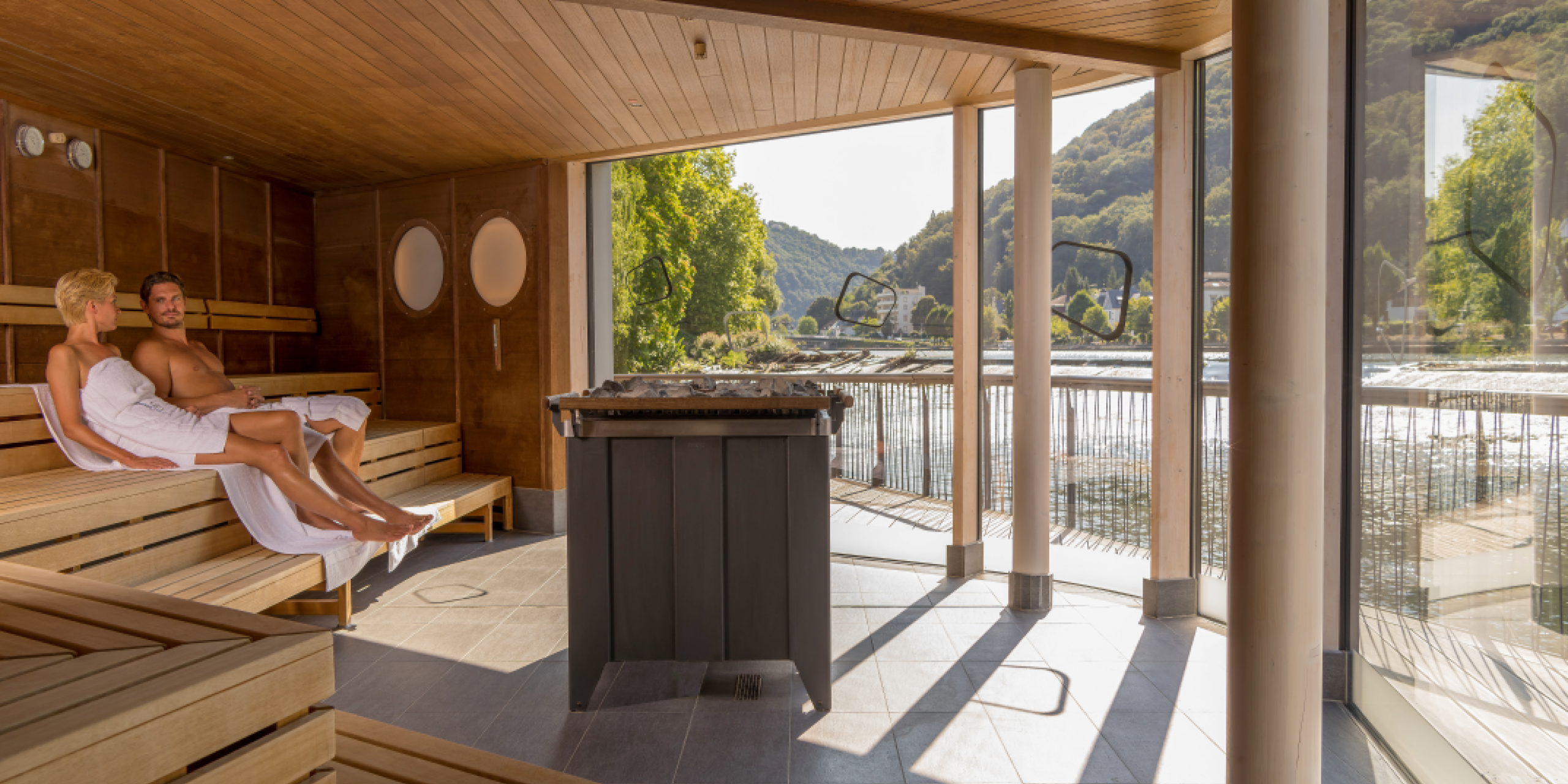 Ein modernes Spa mit Holzverkleidung. Ein Paar sitzt entspannt in Badehandtüchern auf Holzliegen, während große Fenster den Blick auf eine grüne Landschaft und einen Fluss bieten.