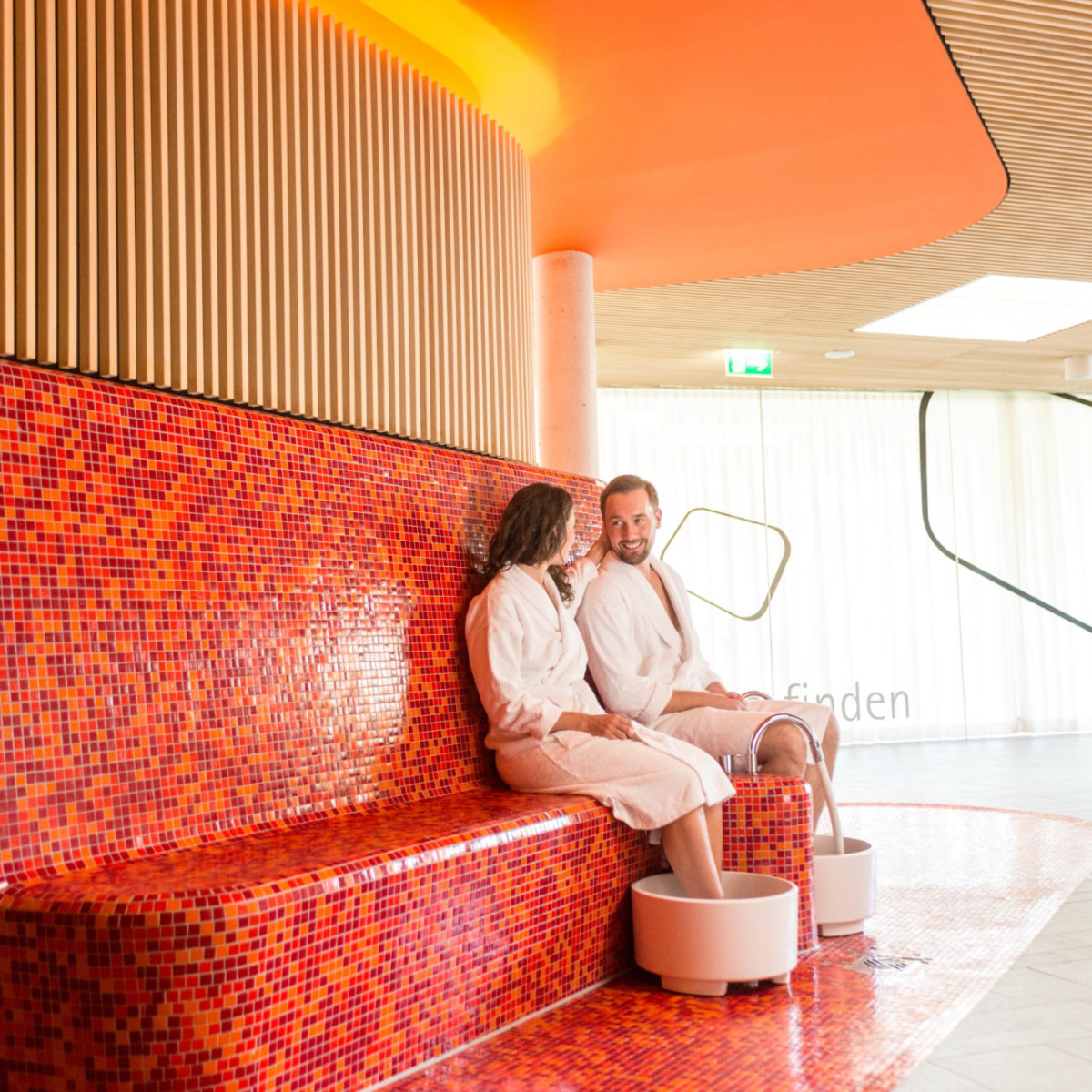 Ein Paar in Bademänteln sitzt auf einer rot gefliesten Bank in einem modernen Spa. Sie genießen die entspannende Atmosphäre und plaudern entspannt, während ihre Füße in Wannen mit Wasser ruhen.