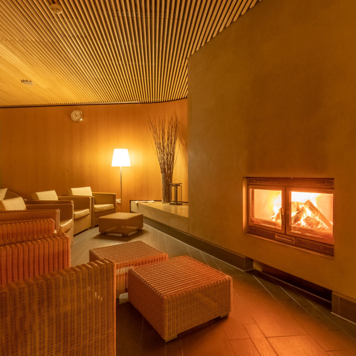 Gemütlicher Wellnessbereich mit modernen Rattanmöbeln, warmem Licht und einem Kamin. Helle Wände und Holzdecke schaffen eine entspannte Atmosphäre für Erholung und Entspannung.