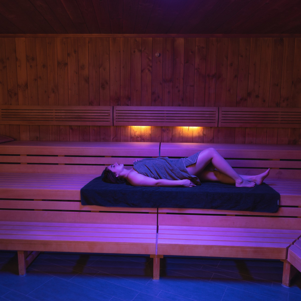 Eine entspannte Person in einem Handtuch liegt auf einer Holzbank in einer Sauna, umgeben von warmem, sanftem Licht in violetten und goldenen Tönen. Die Atmosphäre strahlt Ruhe aus.