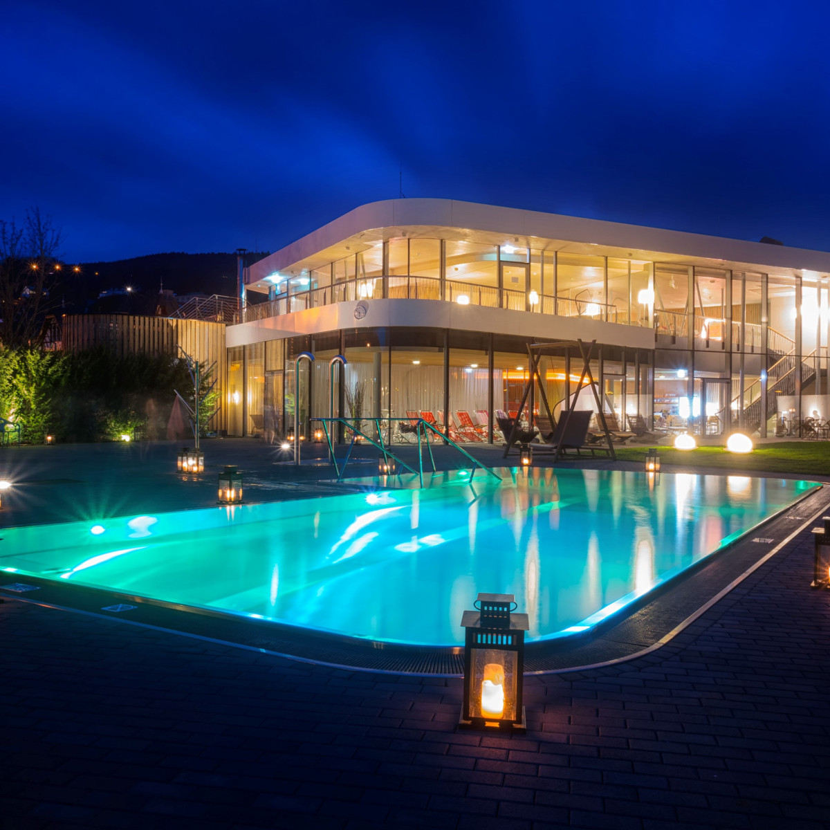 Moderne Villa mit gläserner Fassade und beleuchtetem Pool, umgeben von leuchtenden Laternen. Der Nachthimmel strahlt in tiefem Blau, während das Wasser sanft reflektiert.