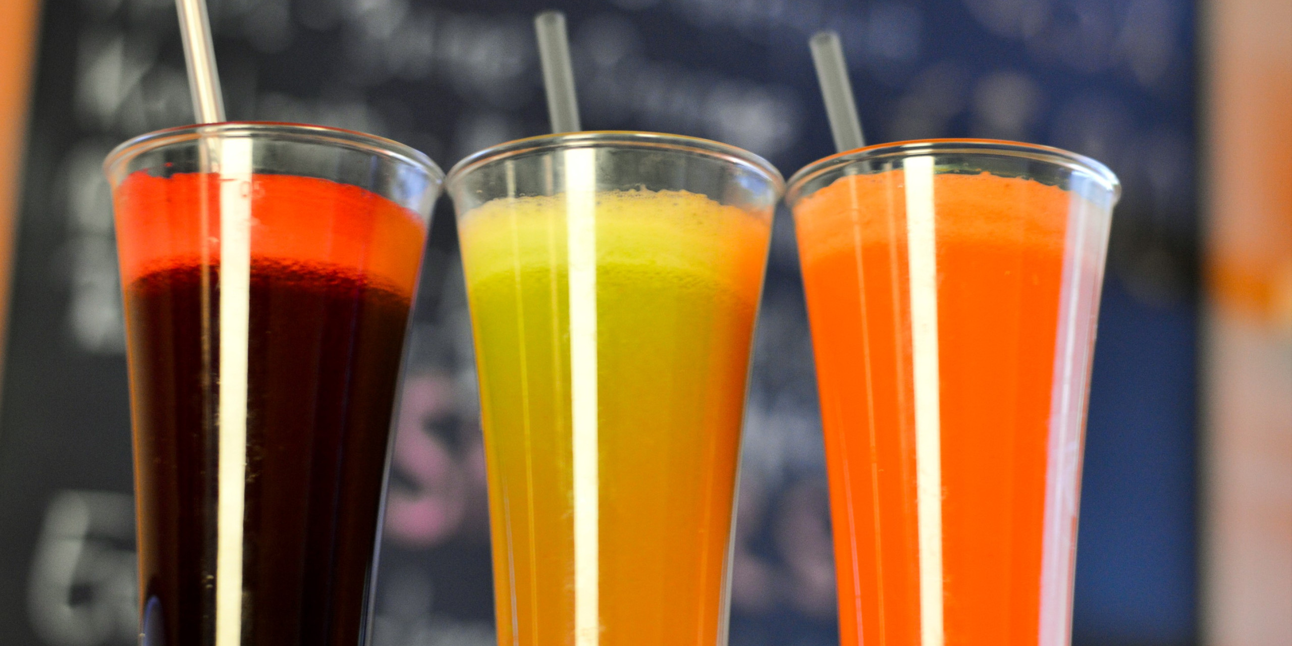Drei hohe Gläser mit bunten Getränken stehen nebeneinander: links ein roter Drink, in der Mitte ein gelb-grüner und rechts ein orangener Drink. Alle haben einen Strohhalm. Der Hintergrund ist verschwo