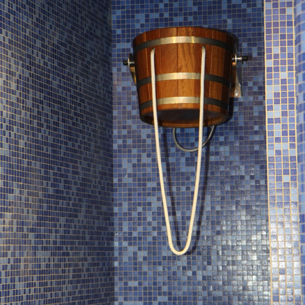 Ein Holzfass mit Alu-Applikationen hängt an einer Wand in einem blauen Mosaik-Bad. Die Wände sind mit kleinen blauen Fliesen bedeckt, die eine entspannende Atmosphäre schaffen.