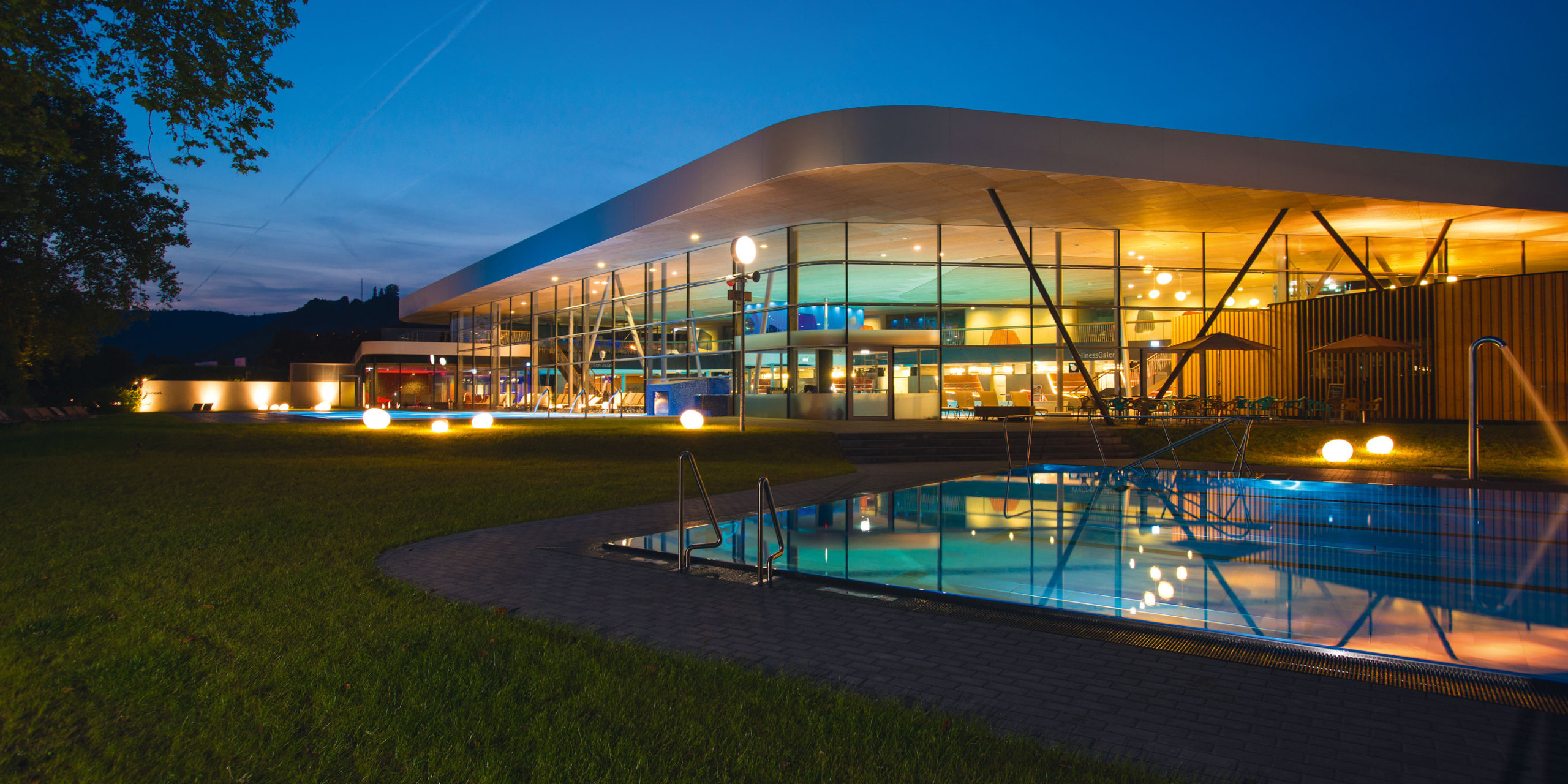 Modernes Gebäude mit großen, verglasten Fenstern bei Abenddämmerung. Umgeben von Grünflächen und einem beleuchteten Pool, dessen Wasser die Lichter reflektiert.