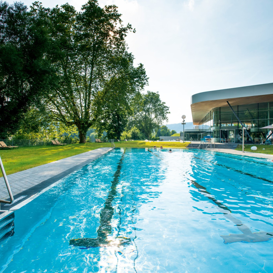 Ein einladender Schwimmbeckenbereich, umgeben von grünen Rasenflächen und Bäumen. Im Hintergrund ist ein modernes Gebäude mit großen Fenstern sichtbar, Sonnenlicht reflektiert auf dem Wasser.