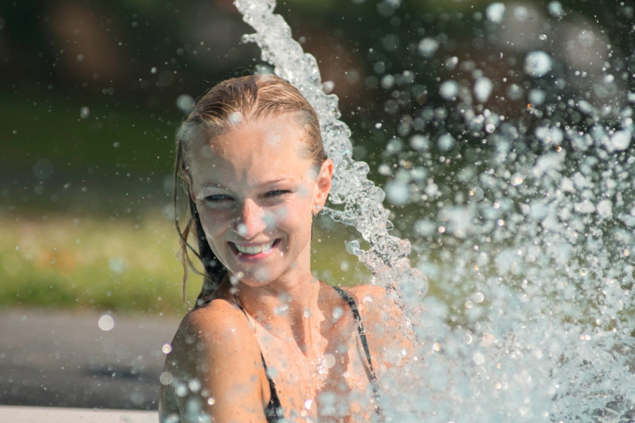 Eine lachende Frau im Pool wird von Wasserstrahlen umgeben. Spritzer fliegen in die Luft, während sie sich fröhlich mit dem Wasser beschäftigt, unter einem sonnigen, grünen Hintergrund.