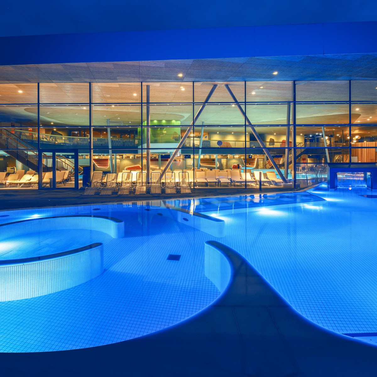 Beleuchteter Pool mit geschwungener Form, umgeben von modernen Liegen. Im Hintergrund sind große Fenster, die eine stilvolle Innenarchitektur und weitere Einrichtungen zeigen. Abendliche Atmosphäre.
