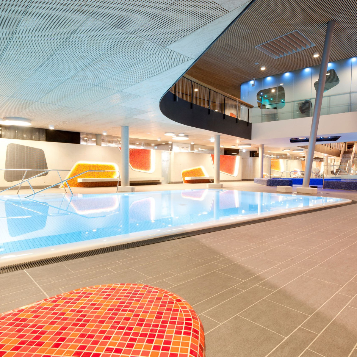 Modernes Schwimmbad mit glasklarem Wasser und farbenfrohen Wänden. Im Hintergrund sind gemütliche Sitzgelegenheiten und ein Obergeschoss mit Glasgeländer sichtbar. Helle, einladende Atmosphäre.