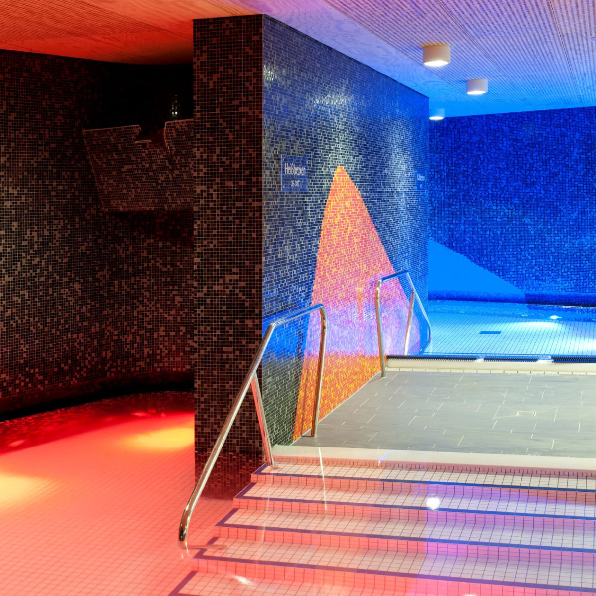 Eine moderne Wellness-Oase mit bunten Mosaikwänden und sanfter Beleuchtung. Treppen führen in einen Poolbereich, umgeben von rotblauem Licht und stilvollen Fliesen. Entspannende Atmosphäre.