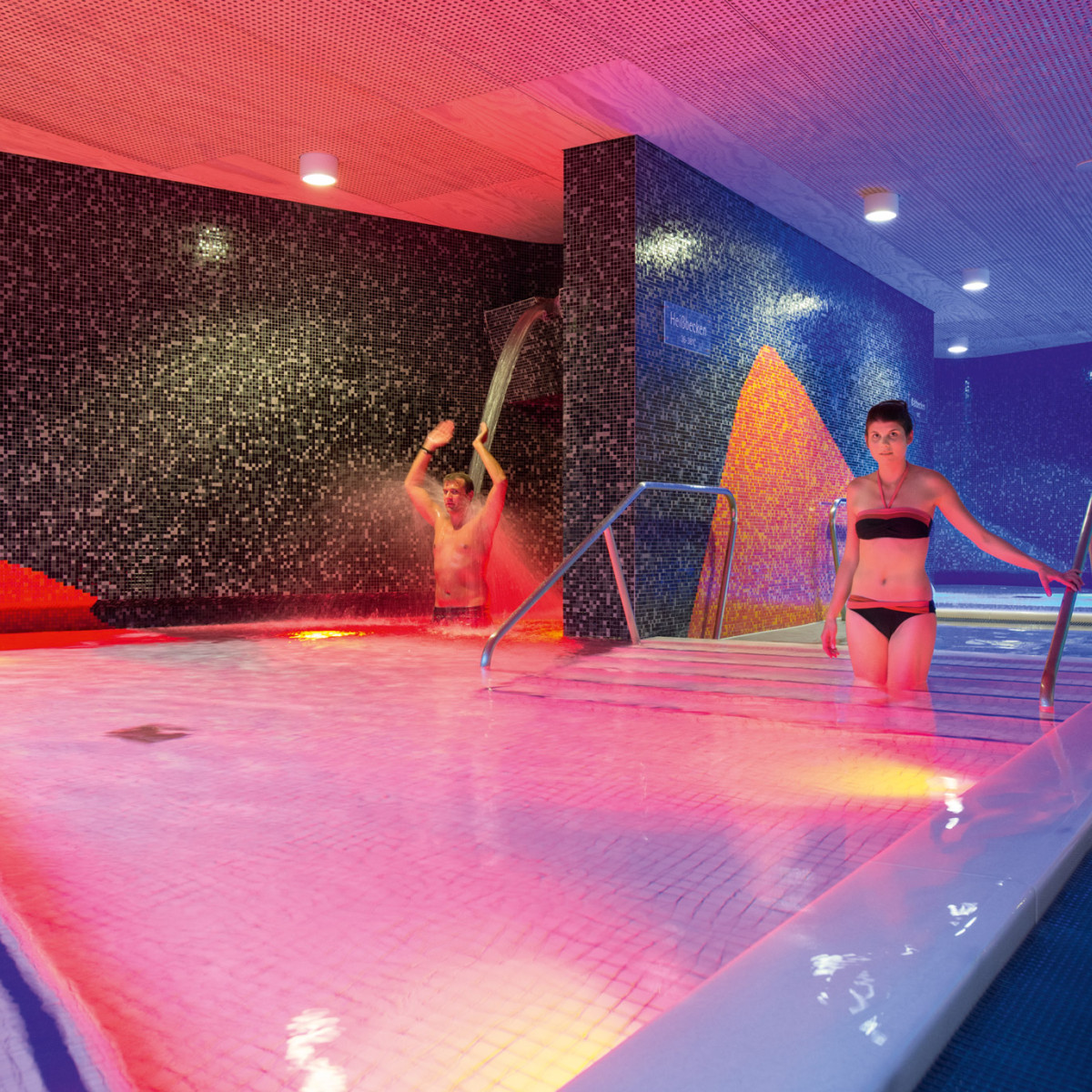 Zwei Personen in einem Spa-Bereich mit bunten Lichtern. Eine Frau steht im Wasser und schaut zur Kamera, während ein Mann im Hintergrund mit Wasser spielt. Mosaikfliesen schmücken die Wände.
