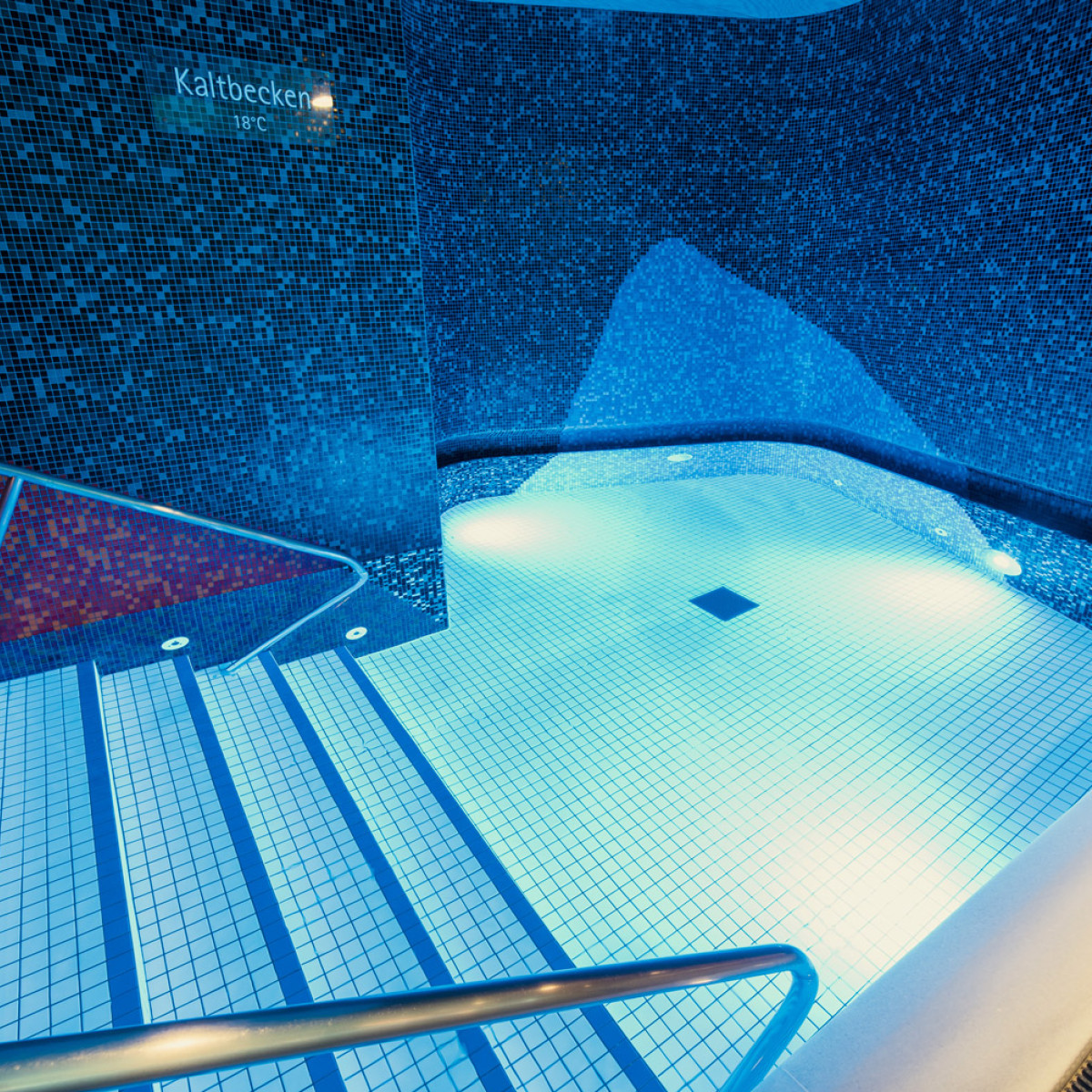 Klares, blaues Kaltbecken mit mosaikverkleideten Wänden. Stufen mit Handlauf führen ins Wasser, das sanft beleuchtet ist. Entspannende Wellness-Atmosphäre in einem modernen Spa-Bereich.