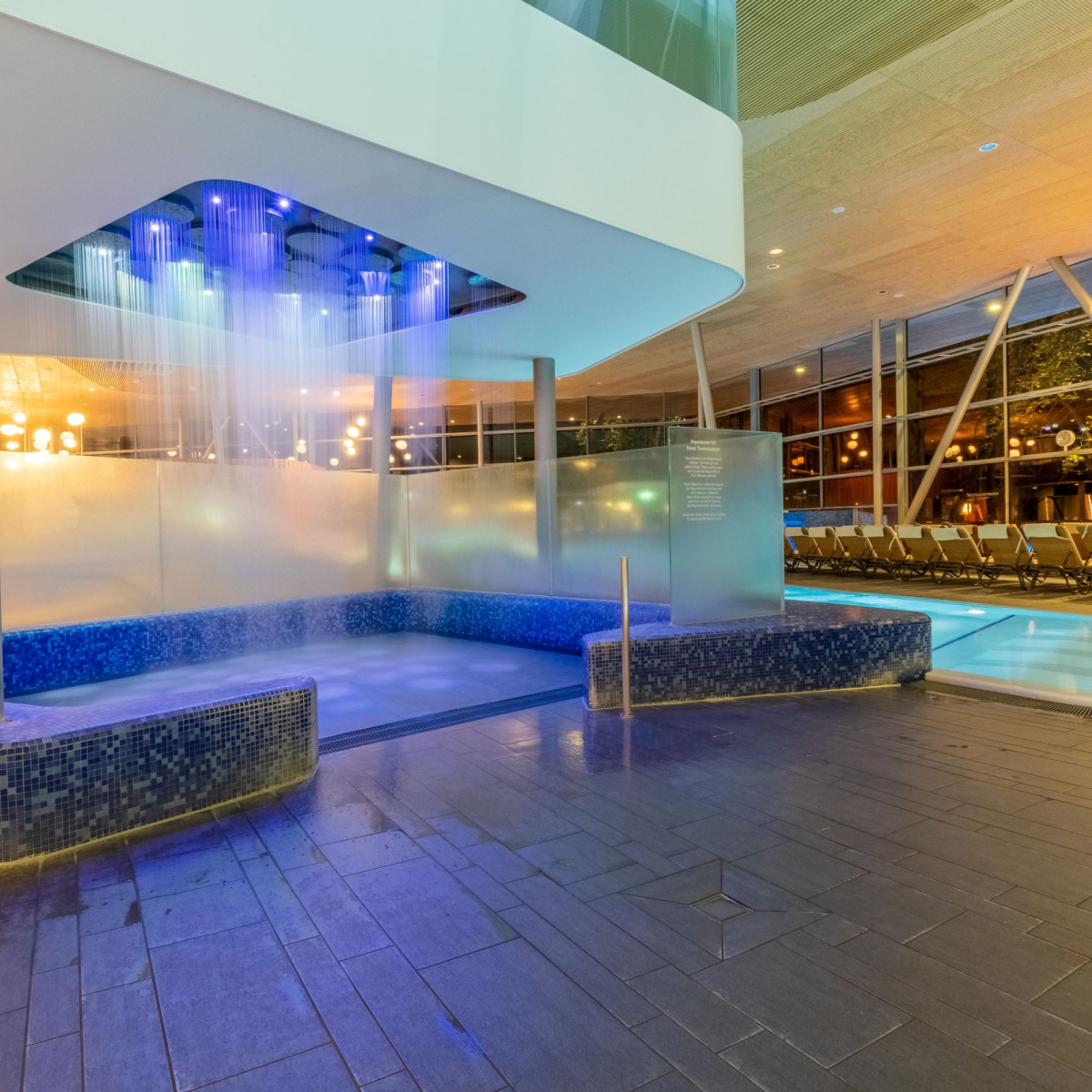 Ein moderner Wellnessbereich mit einem eleganten Jacuzzibereich, blauen Fliesen und einem LED-beleuchteten Wasserfall. Große Fenster bieten Blick auf eine entspannende Außenlandschaft.