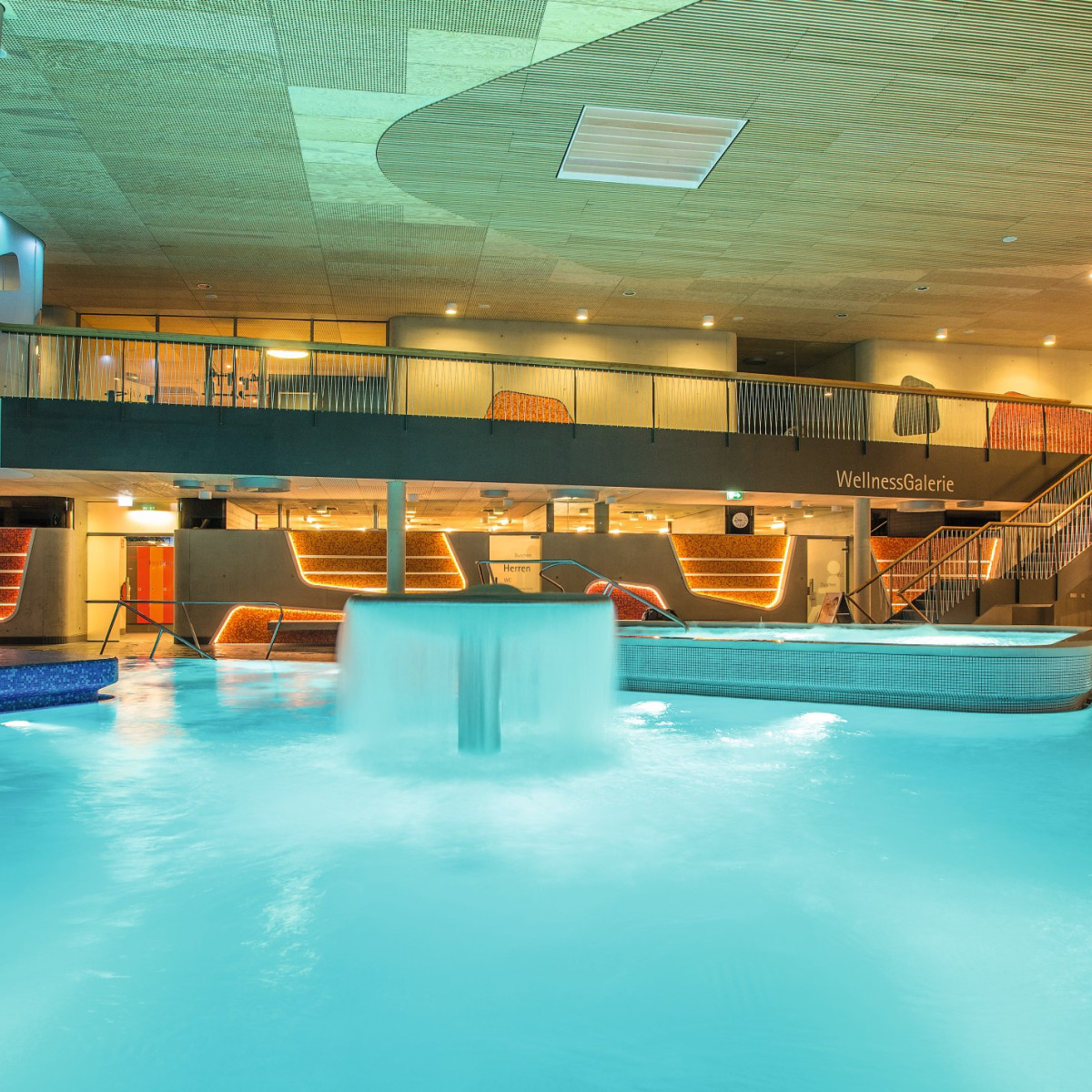 Innenansicht eines modernen Wellnessbereichs mit einem beheizten Pool, einer Wasserfontäne und stilvollem Mobiliar. Warmes Licht und Holzdetails schaffen eine entspannende Atmosphäre.