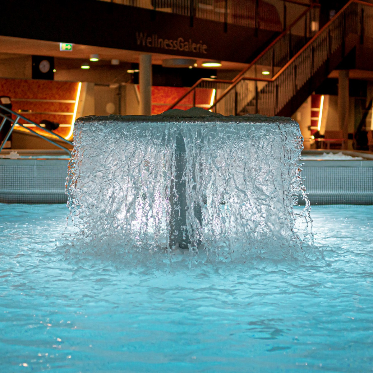 Ein modernes Wellnessbereich mit einem sprudelnden Wasserfall in einem klaren, blauen Pool. Die stilvolle Beleuchtung schafft eine entspannende Atmosphäre, während im Hintergrund ein gemütlicher Sitzb