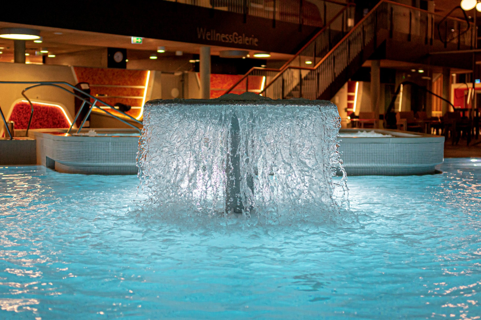 Ein modernes Wellnessbereich mit einem sprudelnden Wasserfall in einem klaren, blauen Pool. Die stilvolle Beleuchtung schafft eine entspannende Atmosphäre, während im Hintergrund ein gemütlicher Sitzbereich sichtbar ist.