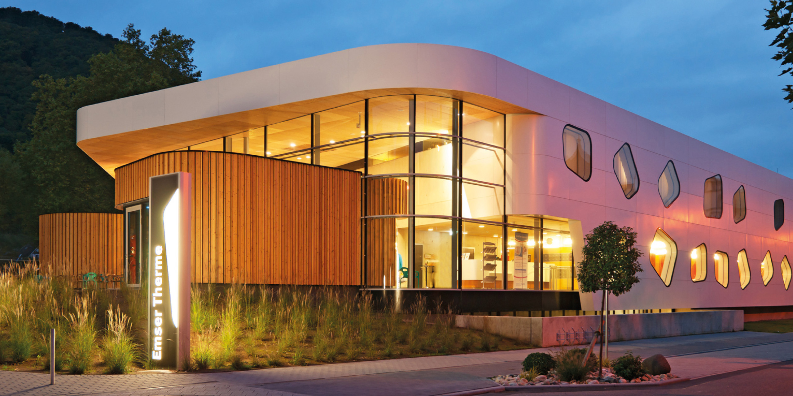 Moderne Wellness-Einrichtung mit markanter Architektur. Die Fassade kombiniert Holz und Glas, während besondere Fensterformen das Gebäude hervorheben. Weiches Licht in der Dämmerung schafft eine einladende Atmosphäre.
