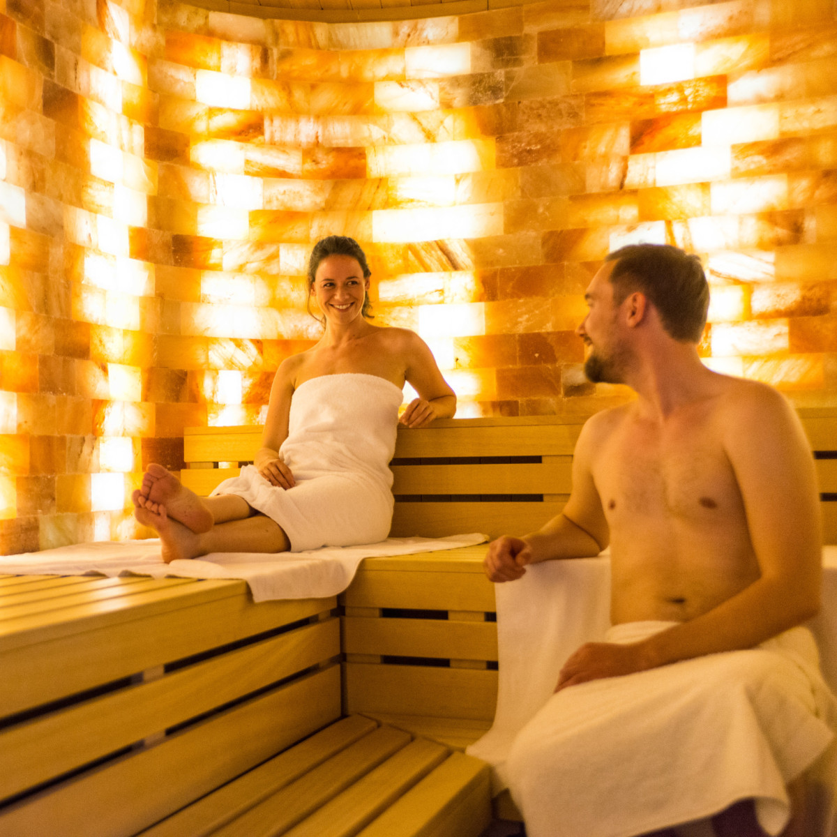 Ein entspannendes Sauna-Ambiente mit goldenem Salzkristall-Wänden. Eine Frau sitzt entspannt auf der Bank, während ein Mann sie anlächelt. Beide tragen Handtücher und genießen die wohlige Atmosphäre.