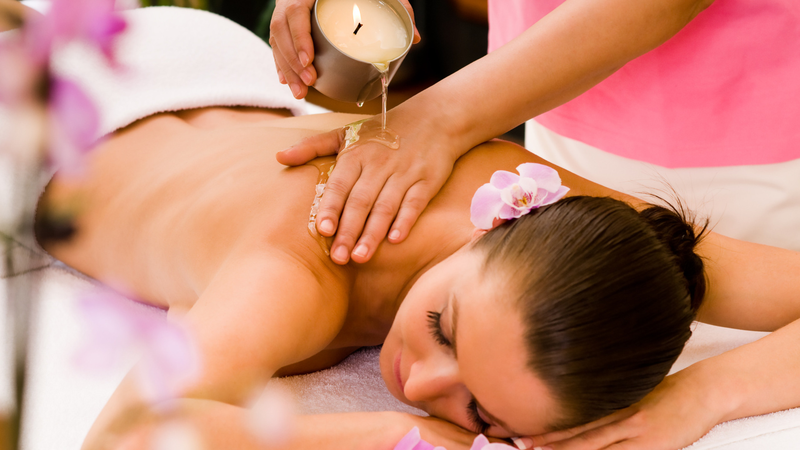Eine Frau liegt entspannt auf einer Liege, während eine Masseurin warmes Öl aus einer Kerze auf ihren Rücken gießt. Violette Orchideenblüten umgeben die Szene und verstärken die entspannende Atmosphär