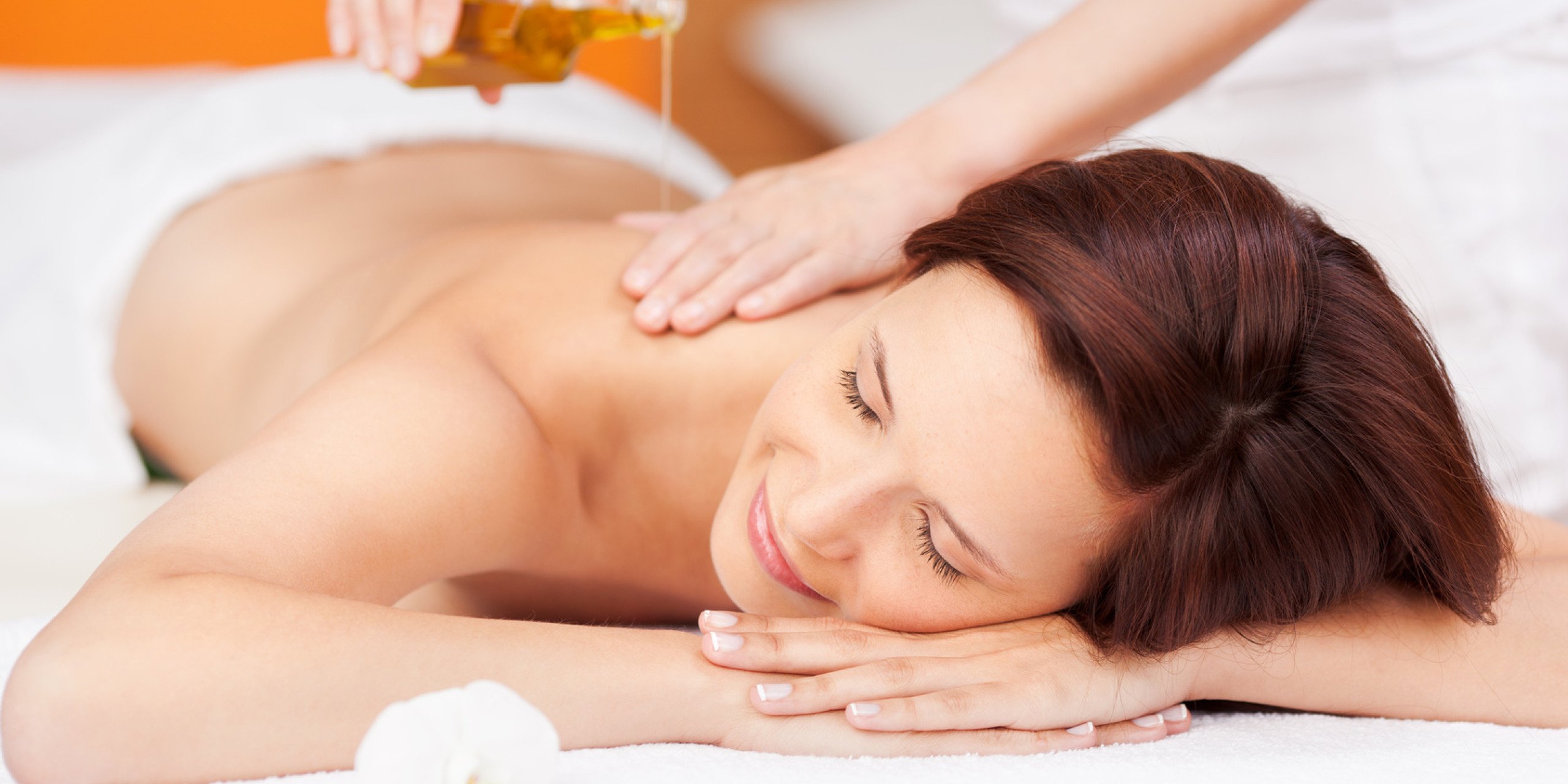 Eine entspannte Frau liegt auf einer Liege, während sie eine Rückenmassage erhält. Eine Therapeutenhand gießt Öl auf ihren Rücken. Eine Blüte liegt neben ihr auf dem Tuch.