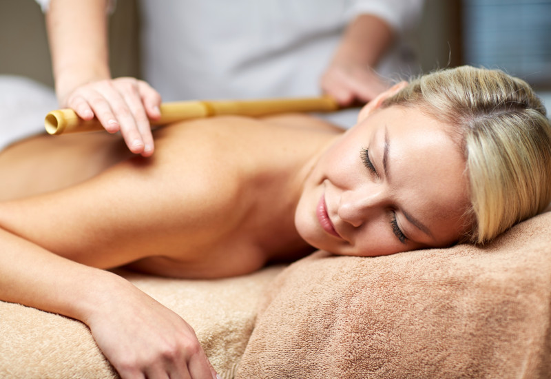 Eine Frau liegt entspannt auf einer Liege, während eine Therapeutin eine Massage mit einem Bambusstab durchführt. Die Atmosphäre wirkt beruhigend und wohltuend, mit sanften, warmen Farben.
