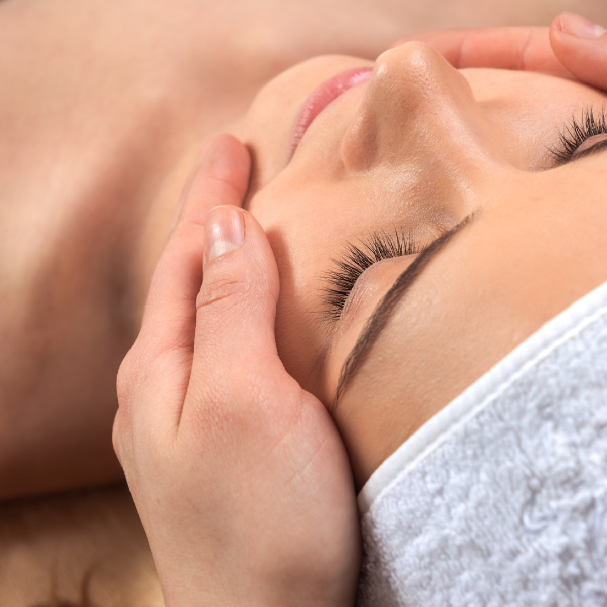 Eine entspannte Frau liegt mit geschlossenen Augen auf einer Liege. Ihre Gesichtshaut wird sanft von einer Hand massiert, während sie ein Handtuch um den Kopf trägt. Die Atmosphäre ist beruhigend und 