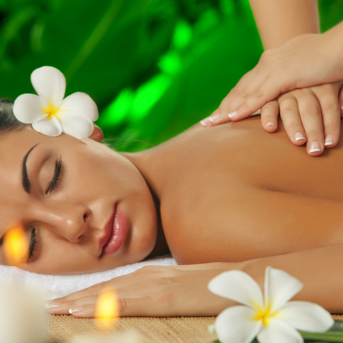 Eine entspannte Frau erhält eine Rückenmassage in einer beruhigenden Umgebung, umgeben von grünen Pflanzen und dekorativen Kerzen. Eine weiße Blume ziert ihr Haar.