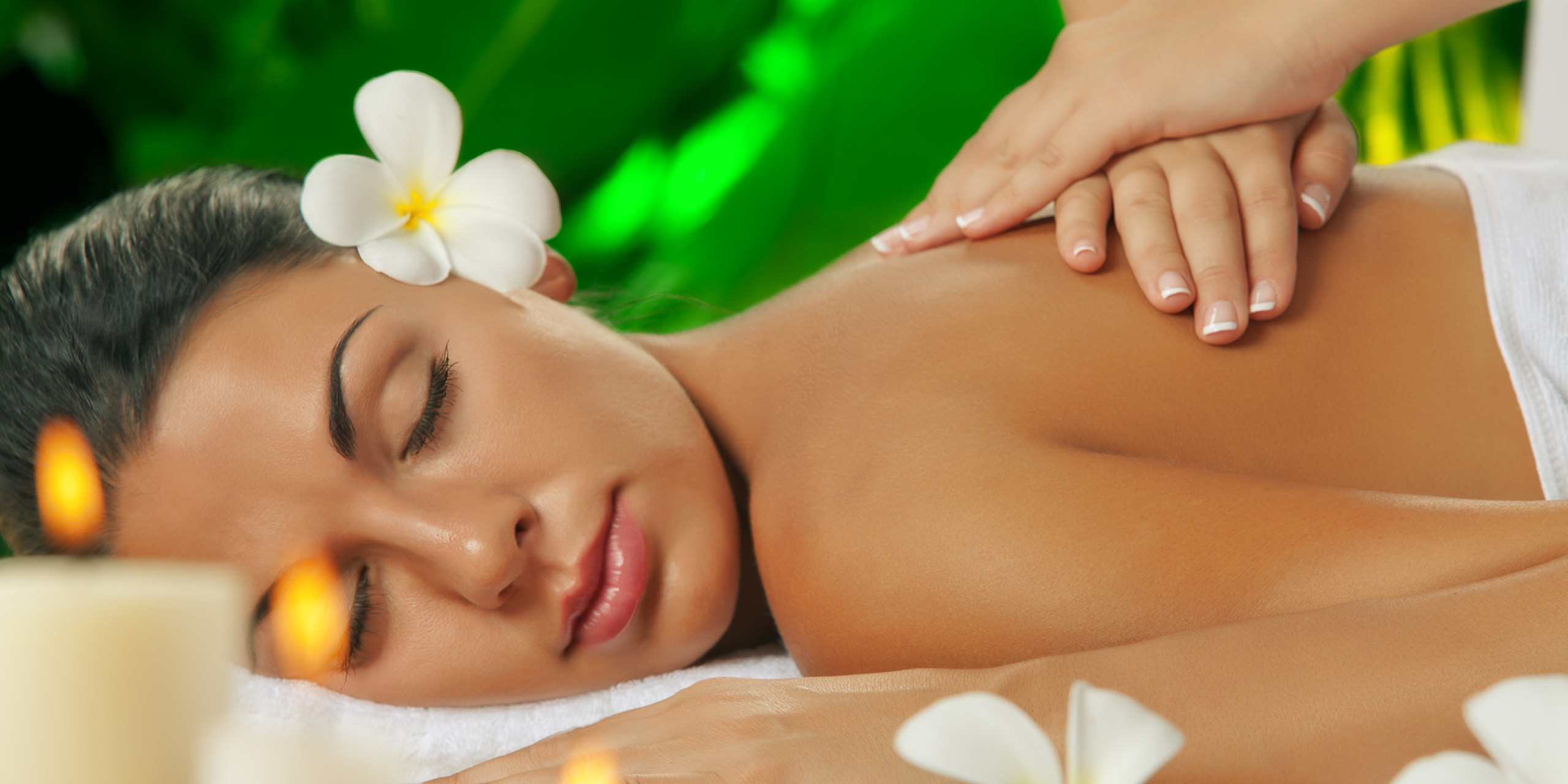 Eine entspannte Frau erhält eine Rückenmassage in einer beruhigenden Umgebung, umgeben von grünen Pflanzen und dekorativen Kerzen. Eine weiße Blume ziert ihr Haar.