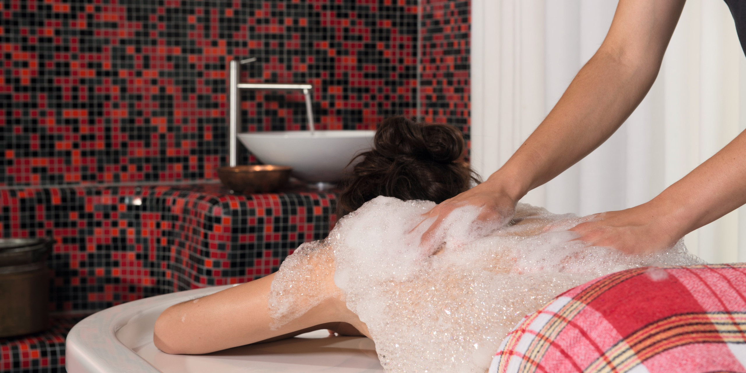 Eine Person liegt entspannt auf einer Badewanne, während eine andere Person sanft ihren Rücken mit Seife einreibt. Im Hintergrund sind rote und schwarze Mosaikfliesen zu sehen.