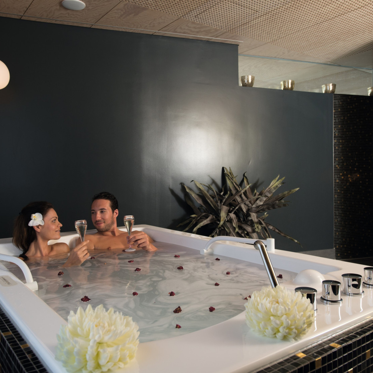 Ein Paar entspannt in einem modernen Whirlpool, umgeben von dunklen Wänden. Sie halten gläserne Drinks und lächeln sich an, während Rosenblätter auf dem Wasser schwimmen. Pflanzen dekorieren den Raum.