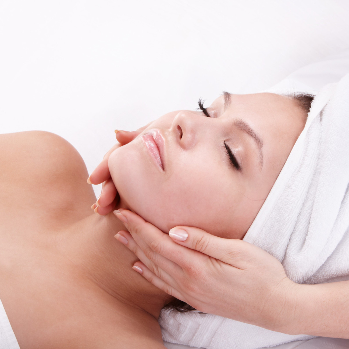Eine entspannte Frau liegt mit geschlossenen Augen auf einer Liege, während eine Hand sanft ihr Gesicht hält. Sie trägt ein Handtuch um den Kopf und ist von einer ruhigen Atmosphäre umgeben.