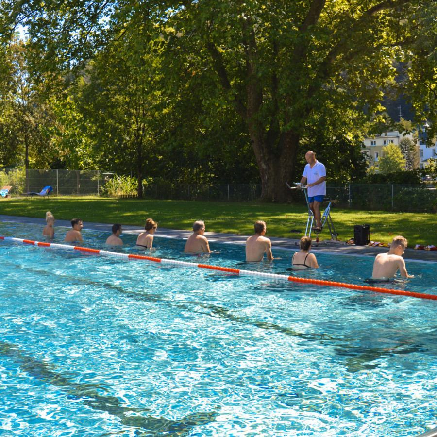 Ein Schwimmbecken an einem sonnigen Tag. Im Vordergrund stehen mehrere Personen im Wasser, während ein Trainer auf einer Leiter Anweisungen gibt. Üppige Bäume umgeben das Becken.