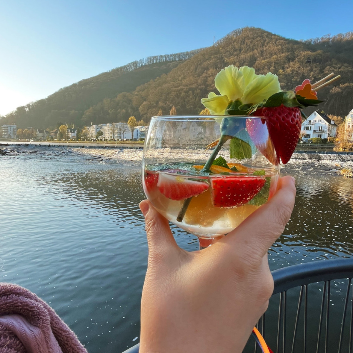 Eine Hand hält ein Glas mit einem fruchtigen Getränk, dekoriert mit Erdbeeren und einer Blüte. Im Hintergrund spiegelt sich die Landschaft im Wasser unter einem klaren Himmel.