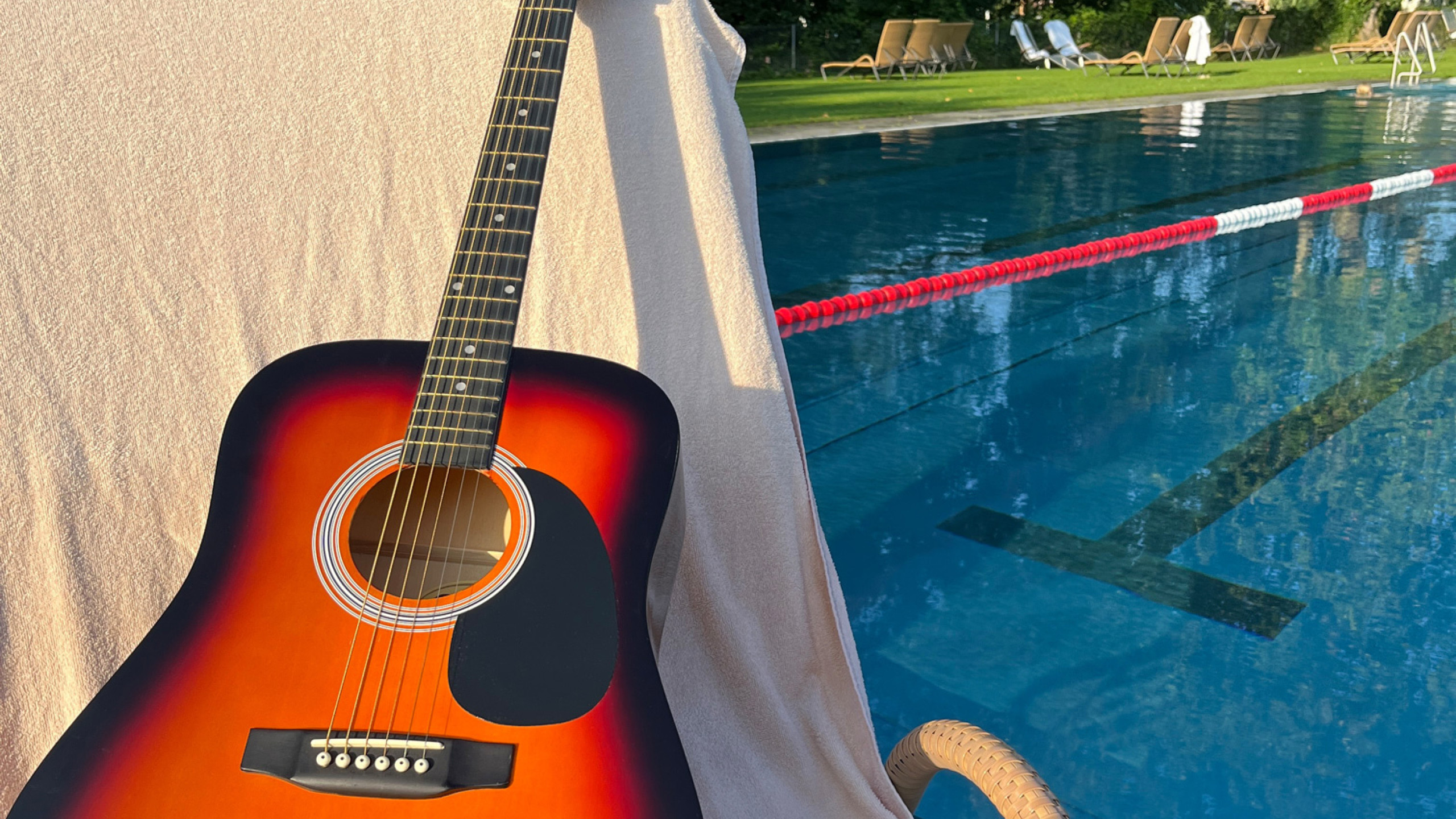 Eine Akustikgitarre in warmen Farbtönen liegt auf einem Handtuch neben einem Swimmingpool. Darunter befinden sich Flip-Flops. Der Pool ist ruhig und von Liegen umgeben.