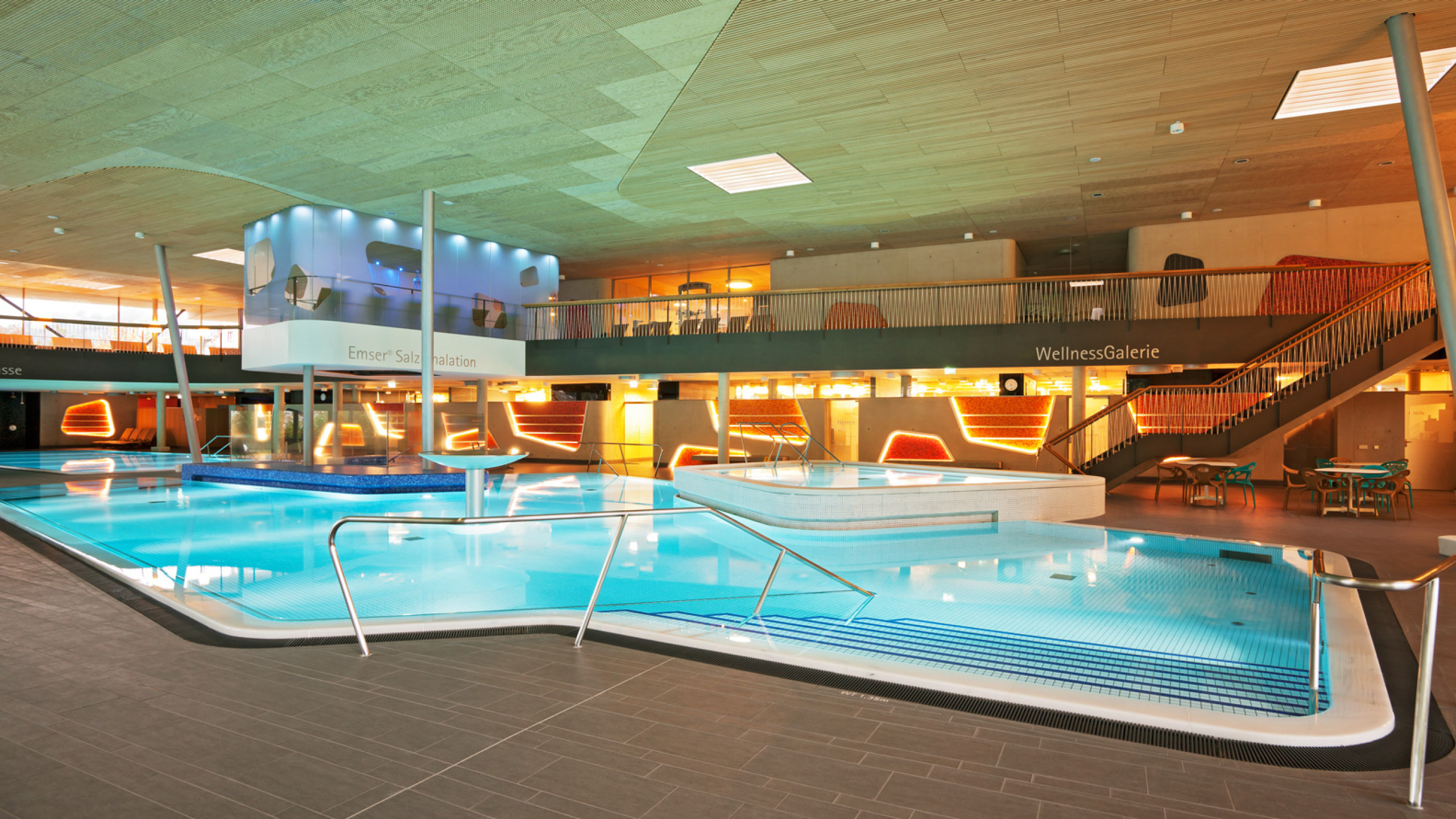 Ein modernes Schwimmbad mit glitschigem Wasser, umgeben von Holz- und Glasarchitektur. Helle Wellnessbereiche und gemütliche Liegen schaffen eine entspannende Atmosphäre.