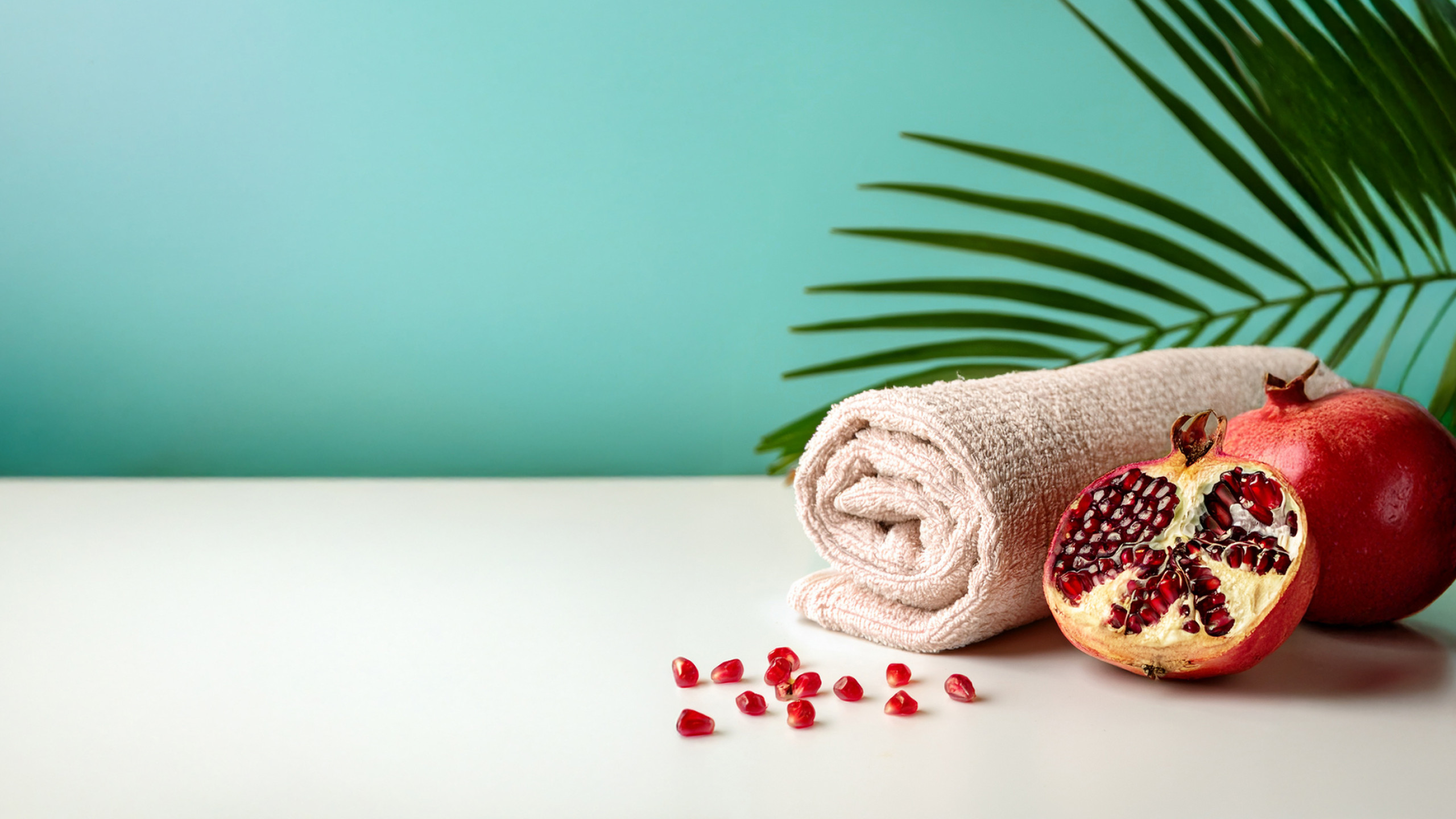 Ein flauschiges, cremefarbenes Handtuch liegt auf einem weißen Tisch, neben einer halb aufgeschnittenen Granatapfel und einer ganzen Granate. Im Hintergrund sind grüne Palmblätter und ein sanfter aqua