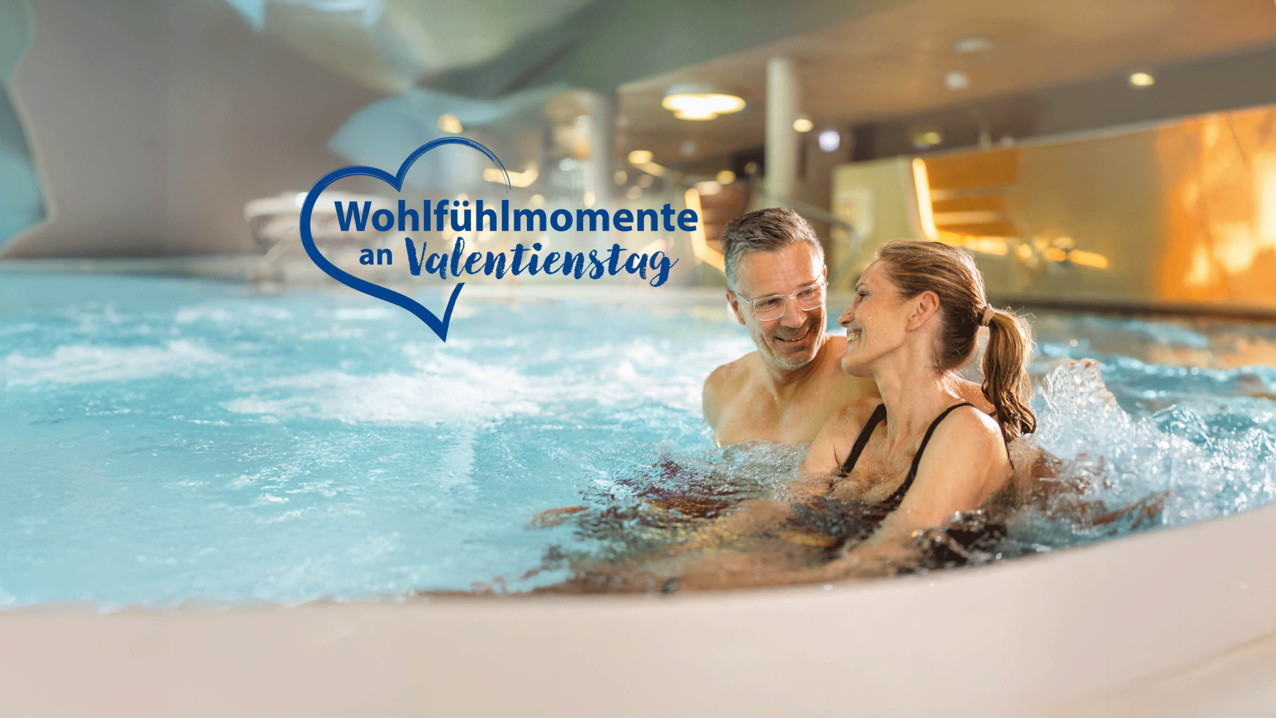 Ein Paar sitzt entspannt in einem Wasserbecken und lächelt sich an. Im Hintergrund sind angenehme Innenräume zu sehen. Text: "Wohlfühlmomente an Valentinstag" in Herzform.